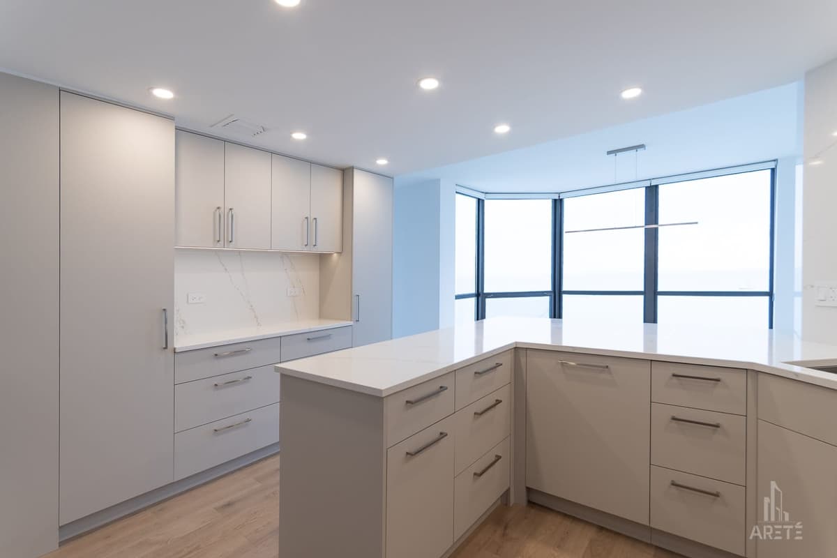 Modern white kitchen with an island, sleek cabinets, and large corner windows letting in natural light.