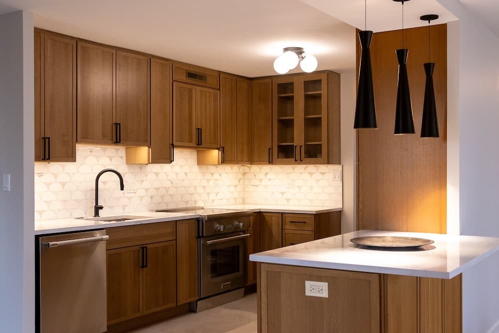 A modern kitchen with wood cabinets, white countertops, and pendant lights. A unique tiled backsplash enhances the design.